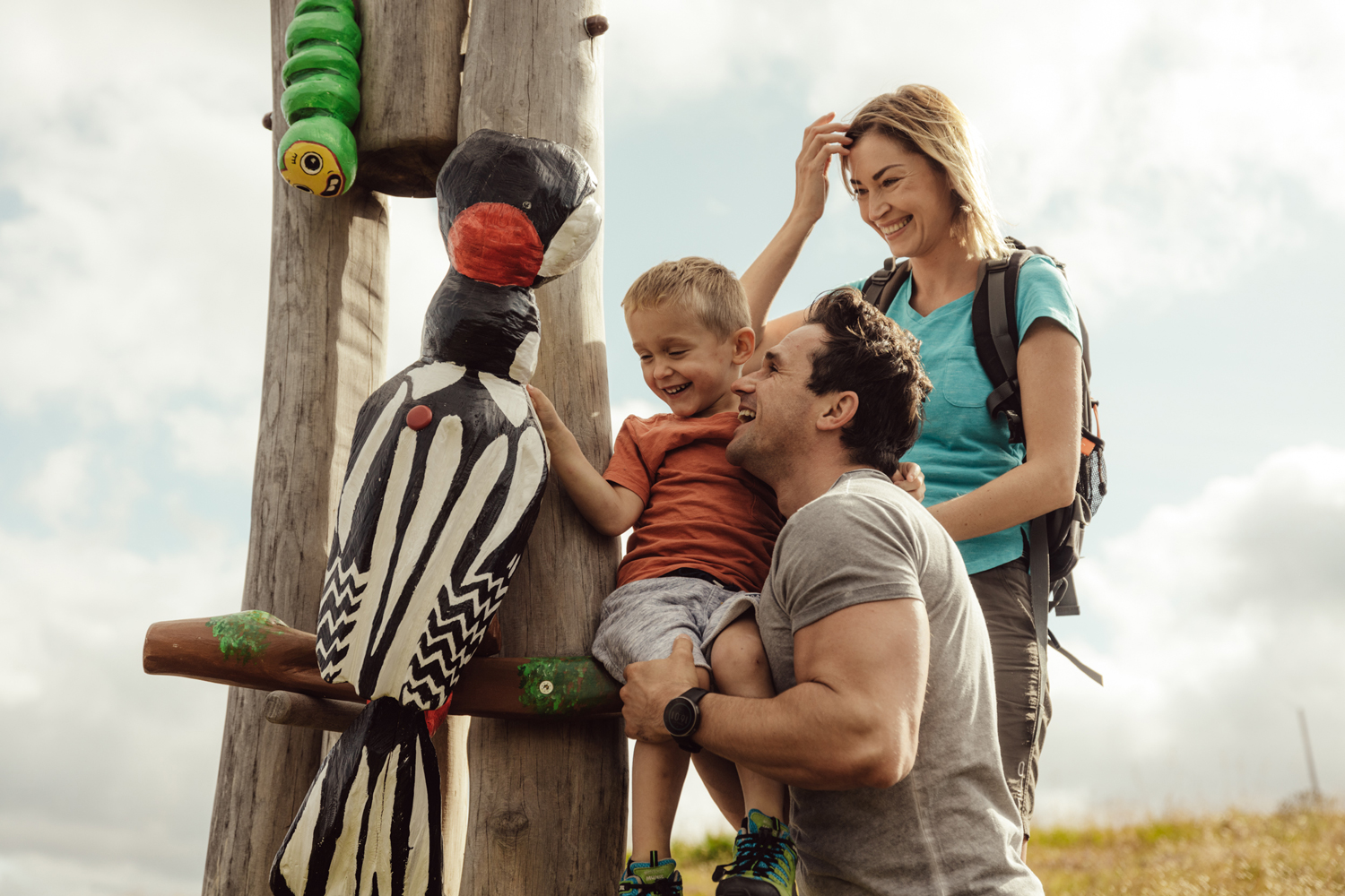 Eine Mutter, ein Vater und ihr kleiner Sohn erkunden einen Specht aus Holz auf der Kindermeile am Golzentipp in Obertilliach